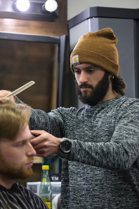 Barber with beard and mustard beanie cuts clients hair in salon with mirror and professional lighting