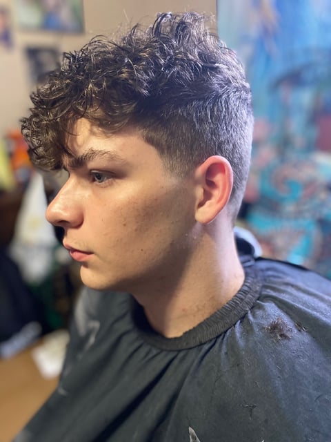 Side profile of a young man with dark curly hair, short fade haircut, wearing a dark shirt indoors